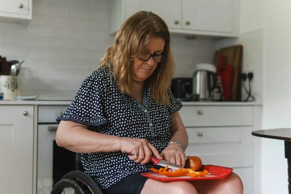 Kvinne i rullestol som skjærer paprika på et skjærebrett på fanget i et lyst kjøkken